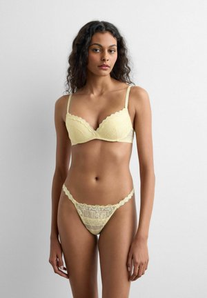 Mujer con el pelo rizado que lleva un sujetador de encaje amarillo pálido y bragas de encaje a juego, de pie frente a un fondo liso de color gris claro.