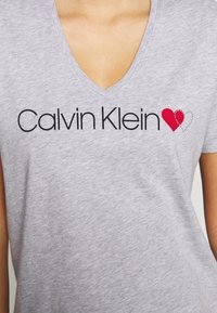 Šedé tričko s výstřihem do V vyrobené z měkké tkaniny, na přední straně má černý nápis "Calvin Klein" s červeným srdcem a srdcem obkresleným v kamíncích.