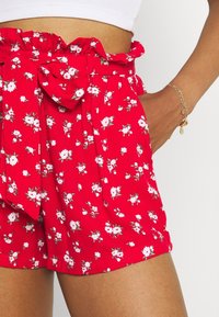 Pantaloni corti rossi con motivi floreali presentano un elastico arricciato in vita, un fiocco annodato, un tessuto leggero e tasche laterali con piccoli motivi di fiori bianchi e verdi.
