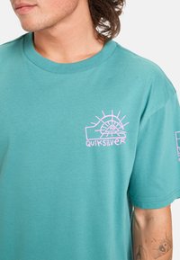 Tyrkysové bavlněné tričko s růžovým grafickým logem se slunečním motivem a názvem značky na levé hrudi. Krátké rukávy a kulatý výstřih.