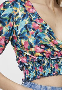 Haut à imprimé floral avec décolleté en V, présentant des couleurs vives de rose, bleu et jaune. Smocks froncés à la taille avec bords volants.
