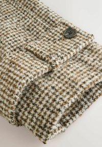 Tissu à motif pied-de-poule brun et crème avec une texture douce, présentant un accent de bouton noir unique sur un bord replié.