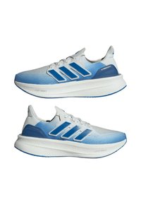 Paire de baskets de course blanches avec un dégradé bleu, bandes Adidas bleues et semelles blanches épaisses portant les mentions « Light Boost » et « Ultraboost ».