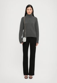 Donna che indossa un maglione grigio a collo alto, pantaloni neri, tacchi aperti neri, e porta una piccola borsa argento con una tracolla dorata.