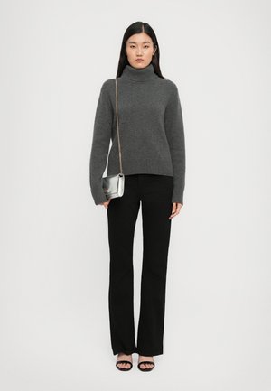Femme portant un pull à col roulé gris, un pantalon noir, des talons ouverts noirs et portant un petit sac à main en argent avec une bandoulière en or.