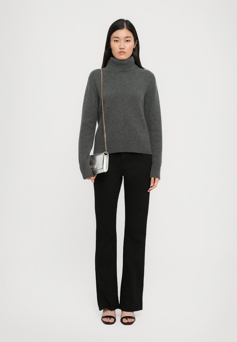 Donna che indossa un maglione grigio a collo alto, pantaloni neri, tacchi aperti neri, e porta una piccola borsa argento con una tracolla dorata.