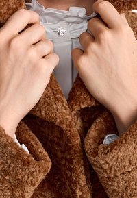 Cappotto marrone soffice con tessuto testurizzato, parzialmente aperto per rivelare una camicia chiara sottostante, caratterizzata da un bottone decorativo.