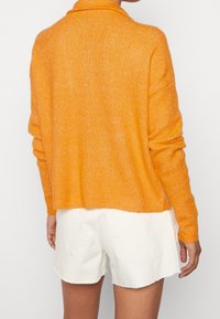 Person wearing a bright orange knit sweater with long sleeves and white denim shorts with frayed hems, shown from the back.