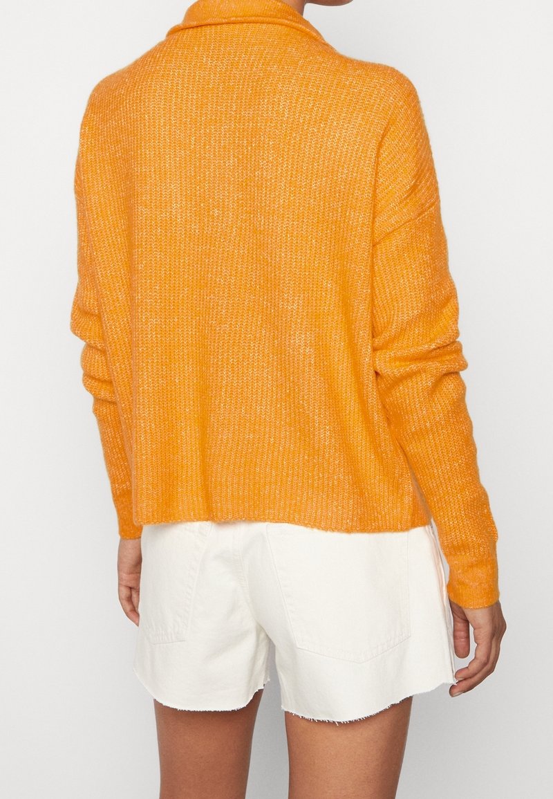 Person wearing a bright orange knit sweater with long sleeves and white denim shorts with frayed hems, shown from the back.