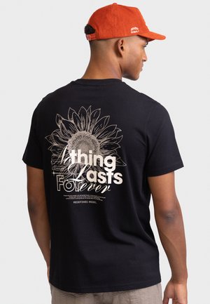 Άνδρας που φοράει μαύρο μπλουζάκι με μεγάλο ηλιοτρόπιο και το κείμενο «Ένα πράγμα διαρκεί για πάντα» στην πλάτη, και πορτοκαλί καπέλο στραμμένο στο πλάι.