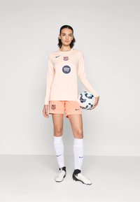 Nike football kit in light pink and orange; long-sleeved shirt with logos and round design, paired with shorts and knee-high white socks.