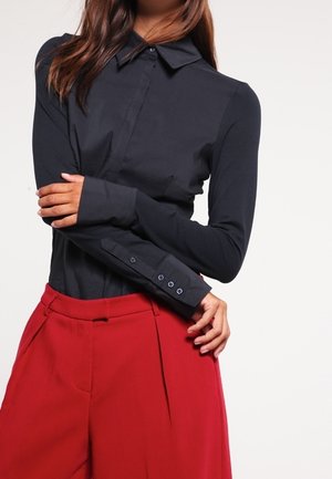 Woman wearing a fitted black long-sleeve button-up shirt tucked into high-waist wide-leg red pants, arms crossed at waist.