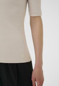 Beige kratký rukáv top s ribkováním, který má přiléhavý střih, měkkou strukturu a malé vyšívané logo blízko lemu. Černé kalhoty jsou částečně viditelné.