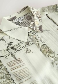 Chemise blanche boutonnée avec imprimé botanique et texte, présentant un col pointu et de subtils plis verticaux sur le tissu.