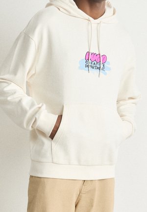 Hoodie couleur crème avec une poche kangourou à l'avant et un capuche avec cordon de serrage. Présente un design graphique en rose et bleu : "DÉPARTEMENT CRÉATIF."
