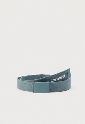 Rullalle kierretty sinivihreä kangasvyö suorakaiteenmuotoisella metallisoljella ja valkoisella "Carhartt"-logolla lähellä päätä.