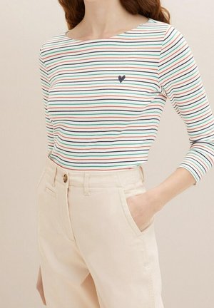 Vrouw draagt een nauwsluitend wit shirt met lange mouwen met dunne, meerkleurige strepen en een klein hartje, gecombineerd met beige broek met hoge taille.