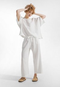 Jonge persoon met krullend haar in losse witte top en broek van textuurstof, barefoot sandalen, poseren met handen op het hoofd tegen een witte achtergrond.