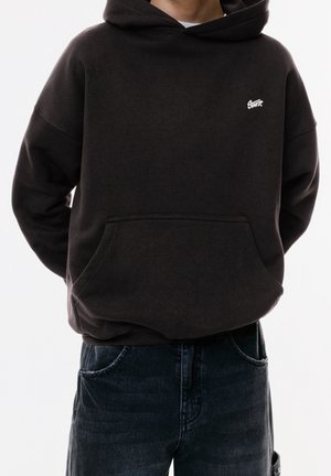 Personne portant un sweat à capuche noir avec un petit logo blanc sur la poitrine et un jean bleu foncé, mains derrière le dos.