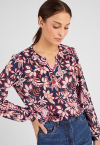 Blusa floreale in navy, con motivi rosa e crema, maniche lunghe, scollo a V leggero e dettagli arricciati sulle spalle.