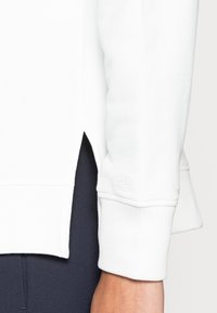Suéter blanco de punto con puños y dobladillo acanalados, con un diseño de abertura lateral. El material parece suave y texturizado, con un detalle de marca mínimo.