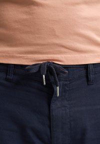 Pantalones azul marino con cintura ajustable mediante cordón, puntas de metal plateadas en el cordón, combinados con una camiseta de color melocotón claro.