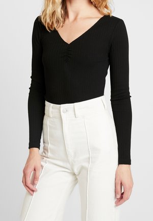 Woman wearing a black ribbed long-sleeve V-neck top tucked into high-waisted white pants with button and zip closure.