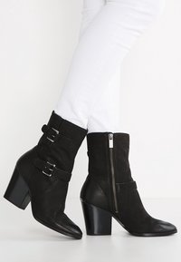 Botines negros de cuero y ante con un acabado texturizado, que incluyen hebillas de metal, cremalleras laterales y tacones de bloque.
