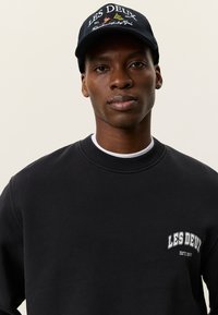 Μαύρο φούτερ με ριμπ γιακά, με λευκό λογότυπο "LES DEUX" στο στήθος. Συνδυάζεται με μπλε σκουφάκι με κεντημένο κείμενο και λογότυπο.