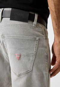 Lichtgrijze spijkerbroek met een driehoekige rode en witte "GUESS" patch op het achterzakje. Het ontwerp heeft klassieke stiknaden en een zwarte riem.