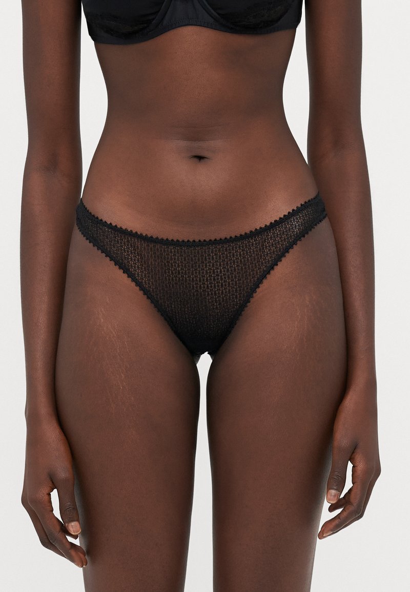 String en dentelle noire avec un motif texturé et des bords festonnés, présentant une taille fine et une couverture minimale. Finition en tissu lisse.