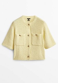 Krótki rękaw żółtego swetra z dzianiny o teksturowanej powierzchni, z dwoma przednimi kieszeniami i okrągłym dekoltem; ma guziki jako zapięcie.