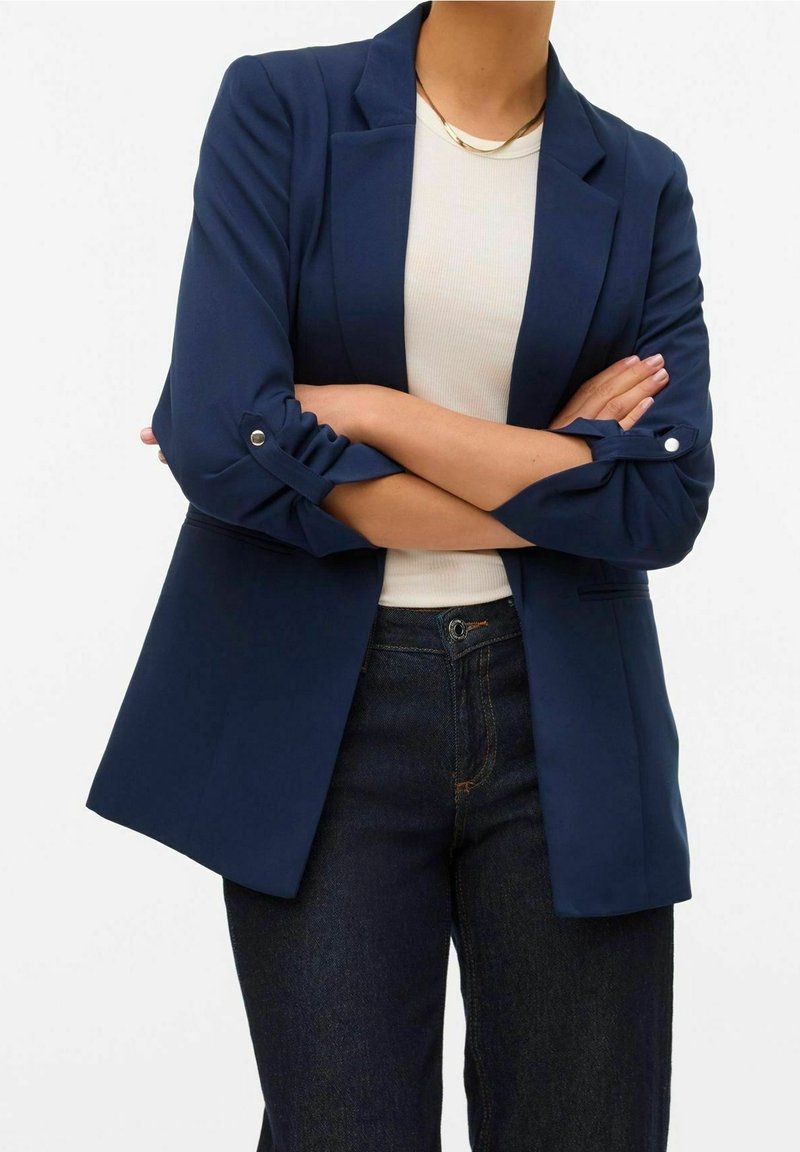 Personne portant un blazer bleu marine avec les manches retroussées sur une chemise blanche et un jean foncé, debout les bras croisés.