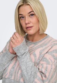 Grijs-roze gebreide trui met een zachte textuur, voorzien van een ronde halslijn en geribbelde manchetten, die gedeeltelijk de handen bedekken.