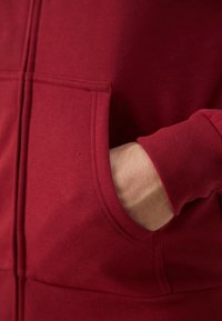 Bordový sveter s kangaroo vreckom, ribbed manžetami a jemnou textúrou látky, s detailným pohľadom na ruku odpočívajúcu vo vrecku.