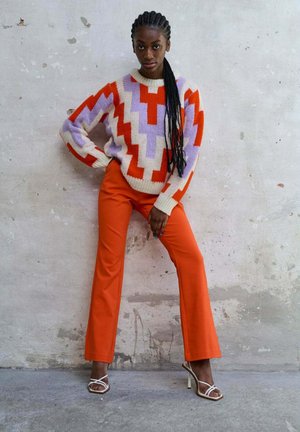 Pull en tricot avec un design en zigzag rouge, violet et crème. Assorti à un pantalon évasé orange et des talons blancs à lanières sur un fond texturé.