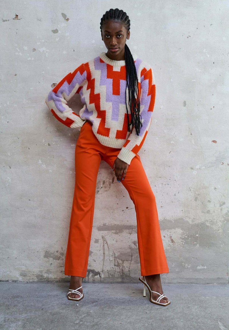 Pull en tricot avec un design en zigzag rouge, violet et crème. Assorti à un pantalon évasé orange et des talons blancs à lanières sur un fond texturé.