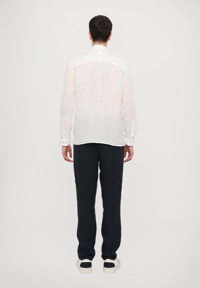White linen shirt with long sleeves, button cuffs, and a subtle gradient. Paired with dark pants and casual shoes, viewed from the back.
