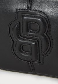 Melnas ādas soma ar izteiktu reljefu logotipu, gludu tekstūru un kontrastējošiem diegiem, demonstrējot elegantu un mūsdienīgu dizainu.