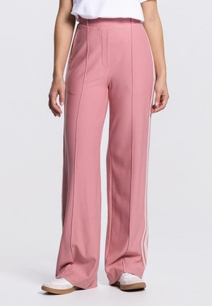 Vrouw draagt hooggetailleerde roze wijde broek met witte strepen aan de zijkanten en witte sneakers, staand tegen een effen achtergrond.