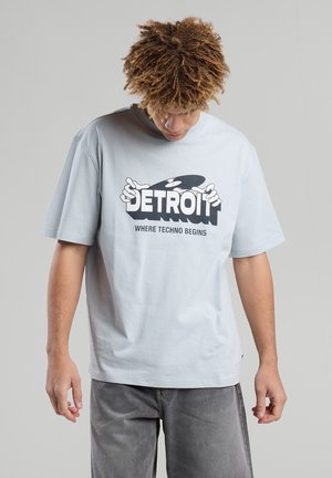 Vaaleansininen ylisuuri T-paita, joka on valmistettu puuvillasta. Paidassa on rohkea musta graafinen teksti "DETROIT" ja "WHERE TECHNO BEGINS" käsikäden kuvioilla.