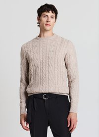 Hellbeiger Strickpullover mit Rundhalsausschnitt, versehen mit strukturierten, erhabenen Mustern und gerippten Bündchen, kombiniert mit dunkler Hose.