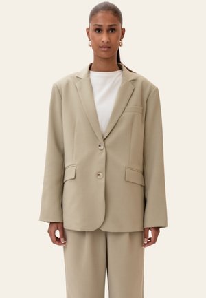 Ženska oblečena v bež preveliko srajčno jakno in ujemajoče hlače z belim topom, stoji pred enobarvnim svetlim ozadjem.