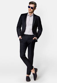 Czarny garnitur z klapami typu notch, jasna koszula w paski z guzikami oraz granatowe zamszowe loafersy. Model nosi okulary przeciwsłoneczne w niebieskim odcieniu.