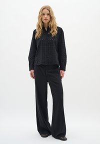 Čierna košeľa s gombíkmi a vyrezávaným vzorom, doplnená širokými čiernymi nohavicami. Hladká textúra a voľný strih celého outfitu.