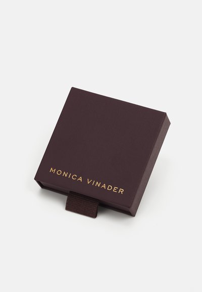 Tumman violetin värinen suorakulmainen korurasia, jossa on teksturoitu pinta. Rasian päällä on kultainen logo "MONICA VINADER" ja ruskea kangasnosto.