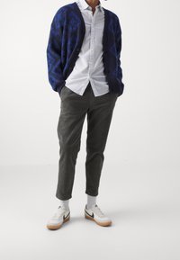 Cardigan a fantasia blu navy, camicia bianca con bottoni, pantaloni in velluto a coste verde oliva e sneakers bianche con dettagli neri. Insieme casual.