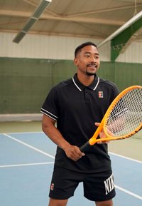 Svart polo skjorte med hvite detaljer, sorte shorts med logo, holder en oransje tennisracket, står på et blått innendørs tennisbane.