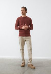 Rozsdabarna kötött pulóver kerek nyakkal, bézs chinos nadrággal és világosbarna slip-on cipővel. Az öltözeten egy kis logó található a pulóveren.
