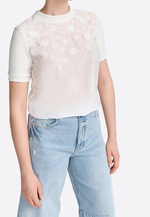 Hvid kortærmet top med gennemsigtigt, broderet blomstermønster og ensfarvede ribkanter, kombineret med lyseblå denimjeans.